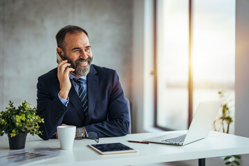 Wall Mural - Mature team leader using his smartphone to text his team mates about new meeting times from his desk in the open office. Young happy Businessman smiling while reading his smartphone. 