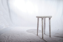 Solitary Wooden Stool In A White Minimalist Setting. Lone, White-painted Wooden Stool Stands Centered On A Textured Surface, Surrounded By Soft White Backdrop