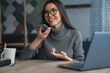 Happy business woman talking on speakerphone sitting at table with laptop on it. Asian female sitting at office having telephonic conversation and smiling
