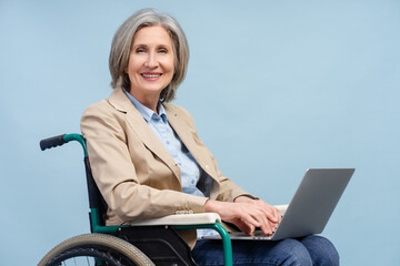 Portrait smiling gray haired senior woman wearing using laptop computer working online sitting in wheelchair isolated on blue background. Freelance job concept