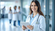 © Kowit - A smiling female doctor holding a tablet stands confidently in a hospital corridor, colleagues in the background.