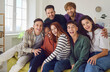 © Studio Romantic - Group of young people sits on a sofa at home together, sharing moments of laughter and happiness. Portrait of a diverse camaraderie of friends, smiling and looking into the camera.
