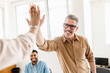 © Vadim Pastuh - A high-five between colleagues, mature man in focus, emphasizes the exuberant success and positive reinforcement prevalent in their modern workspace. The high energy and team-centric atmosphere