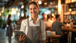 © Kowit - A welcoming waitress in a casual apron smiles as she takes customer orders on a digital tablet in a bustling restaurant.