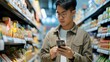 A handsome Asian male walks down the supermarket aisle, browsing shelves and searching for groceries from his mobile phone list, held in his hand.