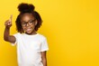little dark-skinned girl, 10 years old, wearing glasses and a white T-shirt, points her finger towards copy space on a yellow background, back to school concept
