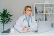 © okrasiuk - Confident mature caucasian senior female doctor in white coat using laptop for telemedicine with patients at hospital. GP make online video call consult patient. Online remote medical appointment.