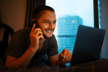 Wall Mural - Young man lying on bed, using smartphone and laptop, evening time. Talking on phone.