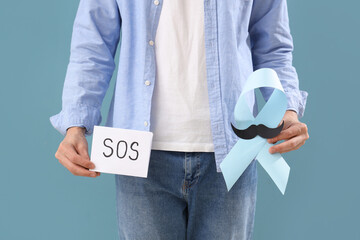 Sticker - Young man holding paper with word SOS and blue ribbon on color background, closeup. Prostate cancer awareness concept