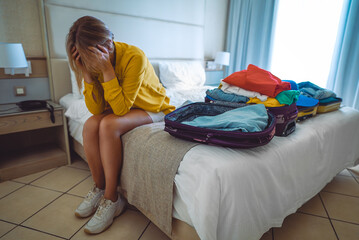 Flight cancellation. Upset woman sitting on bed with suitcases with clothes.