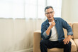 © Chanakon - Handsome asian mature old man sitting on sofa couch in living room at home. Happy Portrait of cheerful smiling senior asian man relax in the house. Mature People and lifestyle