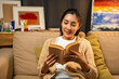 © Chanakon - Asian beautiful woman sitting reading book on cozey couch sofa in living room with warm light at night. People female stay home part time relax at home.