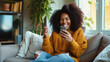 Portrait of young African-American woman celebrating victory, sitting on the couch with a smartphone, raising fist up in triumph, happy black female screams yes