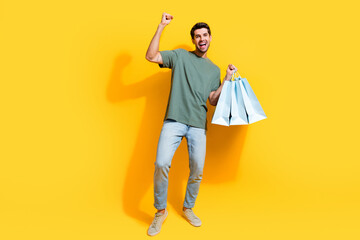 Poster - Full length photo of positive lucky guy wear khaki t-shirt rising fist holding shoppers isolated yellow color background