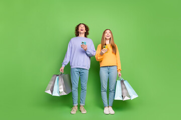 Sticker - Full length photo of two astonished teen buddies hold smart phone mall bags look up empty space isolated on green color background