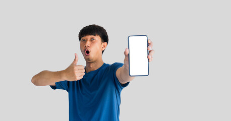 Asian man in blue t-shirt makes wow face and gives thumbs up on mobile phone with blank white smartphone screen isolated on gray background. People recommending new applications or mobile websites
