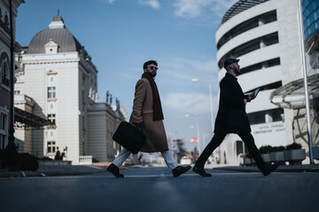 Wall Mural - Confident male professionals in stylish winter attire walking on urban sidewalk, displaying determination and purpose in their stride.