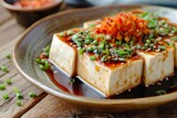 Fototapeta Londyn - Japanese silk tofu with chili ginger and chives served on a ceramic plate over a wooden table with soy sauce topping