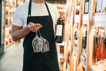 Wall Mural - Two glasses and bottle in hands. Wine shop owner in white shirt and black apron