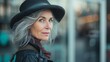 Beautiful stylish good-looking mid-aged woman with grey hair, wearing a black hat, standing on a street in the city