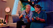 © Raushan_films - Indian Asian Hindu happy smiling old aged cute gen x couple elder older male female man lady sitting cozy sofa couch play video game hold gadget use device enjoy fun joy late night room home house