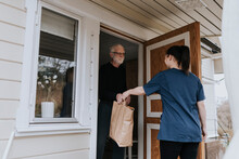 Senior Man Opening Entrance Door To In-home Nurse Who Is Giving Him Paper Bag With Shopping