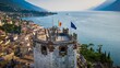 Flags of the EU and Italy on a stone tower in Malcesine with Lake Garda in the background.