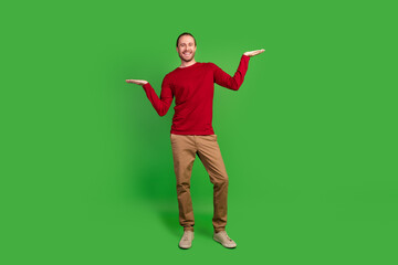 Wall Mural - Full length photo of satisfied positive guy wear red sweatshirt comparing products on arms empty space isolated on green color background