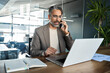 © insta_photos - Busy middle aged business man talking on mobile phone working on laptop sitting at office desk. Mature professional businessman executive making call on cell using computer communicating with client.