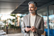 © insta_photos - Happy mature business man executive using mobile cell phone corporate tech outdoors. Smiling handsome middle aged professional businessman holding cellphone looking away standing outside. Copy space