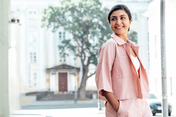 Young beautiful smiling hipster woman in trendy summer pink costume clothes. Carefree female posing in the street at sunny day. Positive model outdoors at sunset. Cheerful and happy