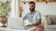 © Studio Nova - man with a beard smiling and waving at the camera, possibly during a video call, in a bright indoor setting with modern decor.