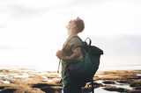 Fototapeta Nowy Jork - Carefree male traveler admiring view of sky near ocean shore with backpack