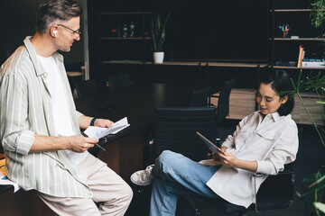 Fototapete - Asian female browsing web content via app on digital tablet during collaborative working process with male colleague in bluetooth earbuds, professional partners discussing business marketing