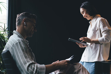 Fototapete - Happy Asian woman using media application on modern touch pad gadget for choosing music playlist for man sitting in chair and analyzing report documents, side view of male and female colleagues