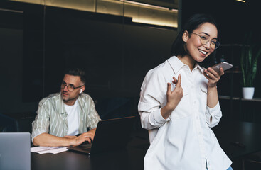 Fototapete - Successful female in classic spectacles for eyes correcting connecting to 4g wireless on smartphone gadget for sending audio sms message during online chatting, happy Asian woman recording sound