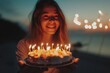 Happy young girl celebrates her birthday in modest intimate atmosphere by the ocean and holds cake with candles.