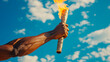 A man's hand holds a torch with the Olympic flame against a blue sky background.