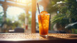 Glass of iced tea with slice of orange and black straw on a table, illuminated by soft sunlight filtering through the foliage.