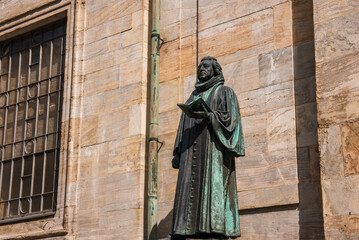 A bronze statue of a historical figure clutching a book stands before a classical building, possibly in Copenhagen, under a clear sky with soft shadows accentuating its form.