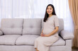 © Issara - Smiling beautiful pregnancy woman sitting on sofa  by the window of her home, showcasing beauty and a positive lifestyle.Asian pregnant female.White background with copy space.Hand on tummy or belly.
