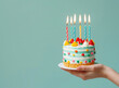 © Artsaba Family - A hand holding a colorful birthday cake with lit candles against a teal background.
