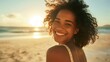 A woman with curly hair smiling at the camera standing on a sandy beach with the sun setting in the background.