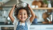 Young child joyfully holding a colander on their head in a kitchen setting.