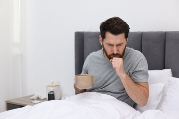 Canvas Print - Sick man with cup of hot drink coughing on bed at home. Cold symptoms