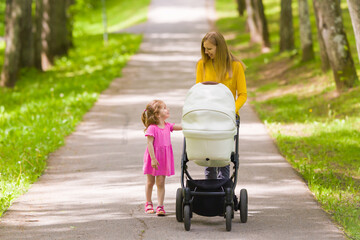 Wall Mural - Happy smiling young adult mother and little daughter pushing white baby stroller and walking at town park in warm sunny summer day. Spending time together. Enjoying stroll. Two child mom. Front view.