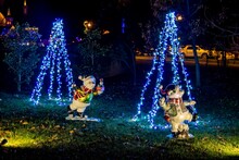 Night Scene Of Decorative Lit Cone-Shaped Christmas Trees With Two Playful Inflatable Snowman Figures In The Foreground, Adding To The Festive Outdoor Decor.