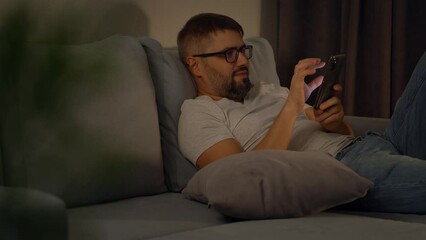 Man holds mobile phone and surfing internet lying on sofa at home in evening. Man rests with phone. sms message, social media, online shopping, search for information