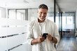© Stock 4 You - Smiling mature Latin or Indian businessman holding smartphone, using cellphone mobile app. Middle aged hispanic employee standing with phone and looking at camera on office background with copy space.