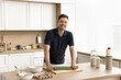 © fizkes - Cheerful handsome baker cooking at home, rolling dough on kitchen table, looking at camera, smiling for portrait. Happy young man baking homemade pastry, enjoying culinary, preparation